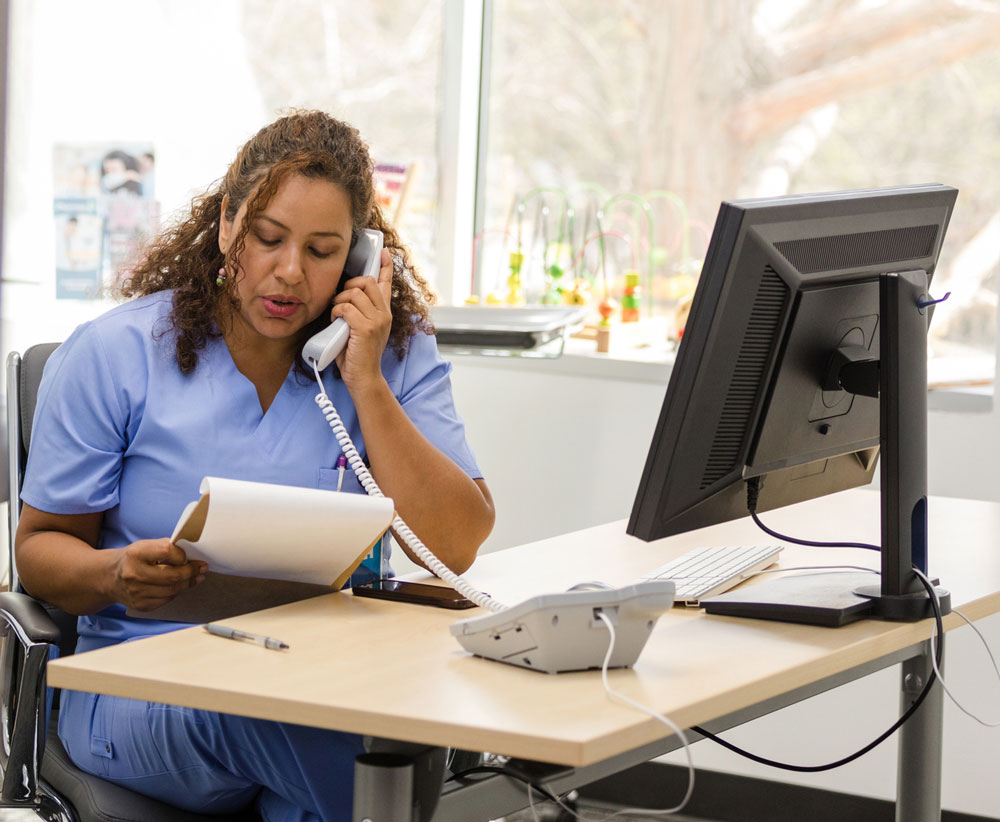 A woman in blue scrubs sits at a desk, talking on a phone while reading from a clipboard. A computer monitor and phone are on the desk, with a window and toys visible in the background.