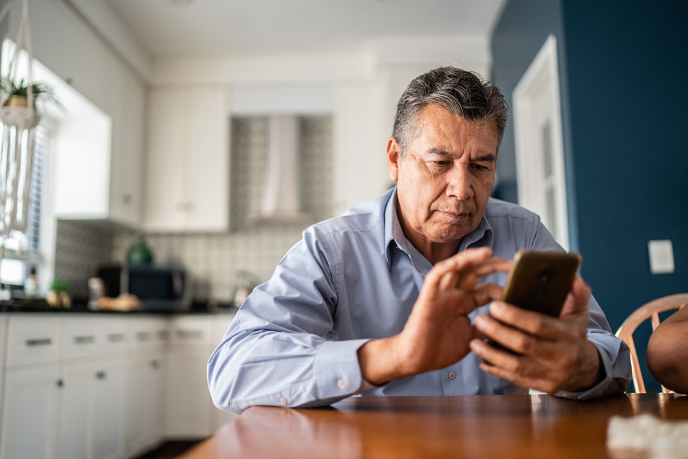 An older man sits at a kitchen table, wearing a light blue shirt, focused on using a smartphone. The background shows a modern kitchen with white cabinets and a blue accent wall.