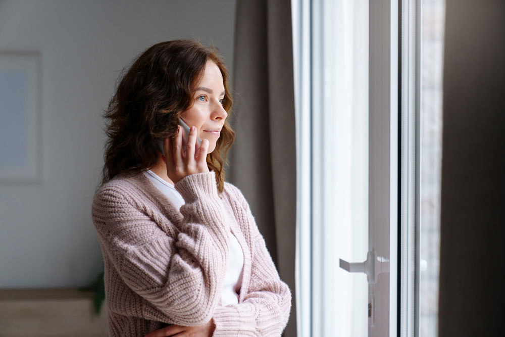 A woman with wavy brown hair, wearing a pink cardigan, stands indoors by a window, looking outside thoughtfully with her hand resting on her cheek.