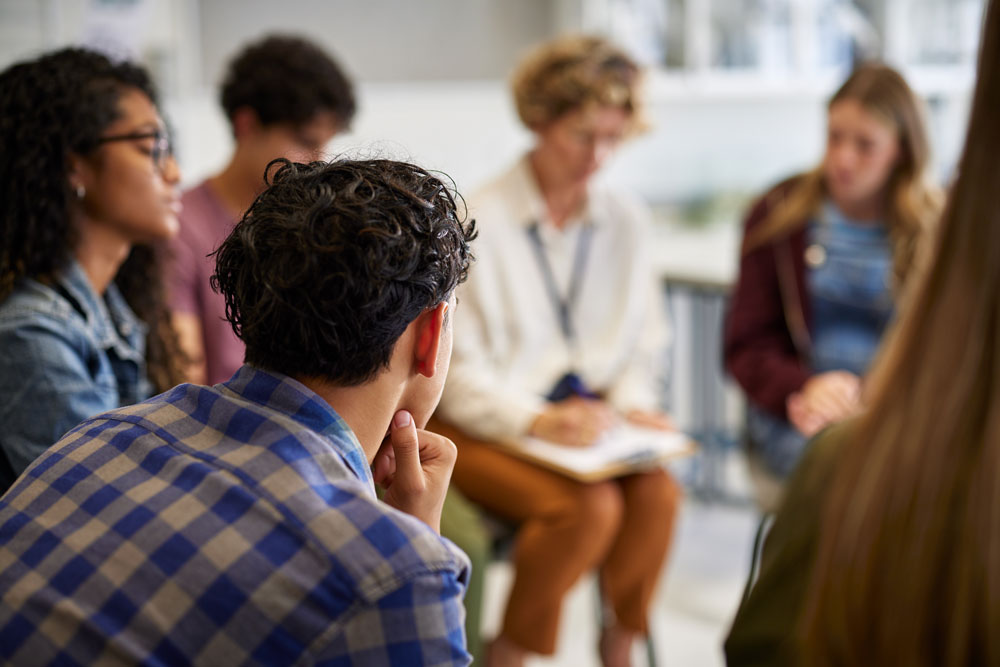 A group of people sit in a circle, engaged in discussion. The focus is on the back of a person with curly hair and a plaid shirt, while others are slightly blurred in the background, suggesting a meeting or support group.