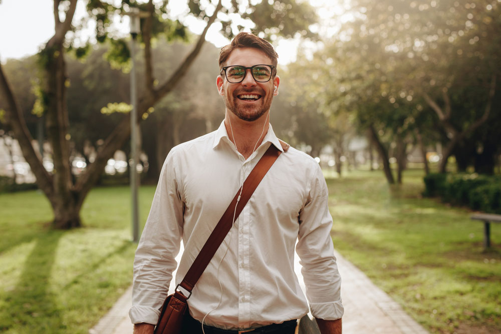 A smiling man with glasses, wearing a white shirt and carrying a shoulder bag, stands on a park path with trees and sunlight in the background.