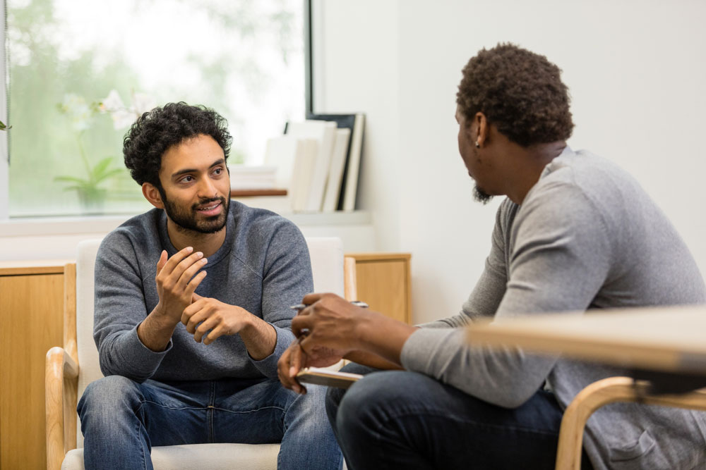 Two men sit facing each other in a bright room, engaged in a conversation. One gestures with his hand while the other listens attentively. They appear relaxed and focused, with books and a window in the background.