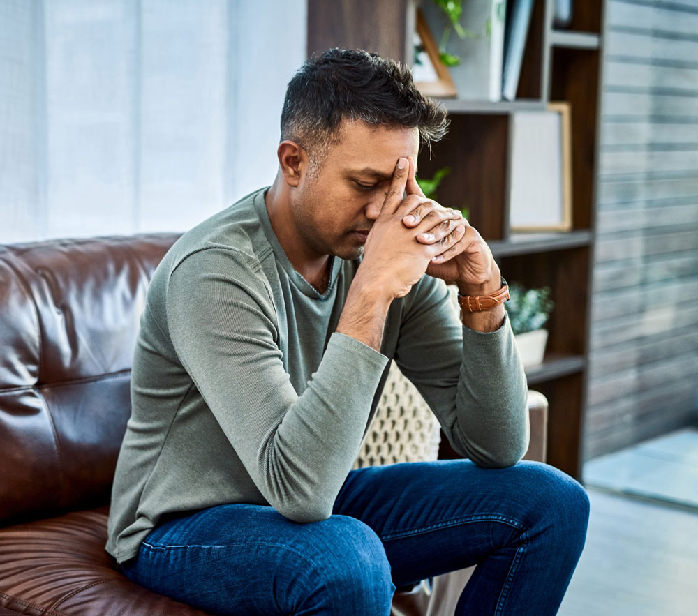 A man sits on a couch leaning forward with his elbows on his knees, eyes closed and fingers touching his forehead, appearing deep in thought or stressed.