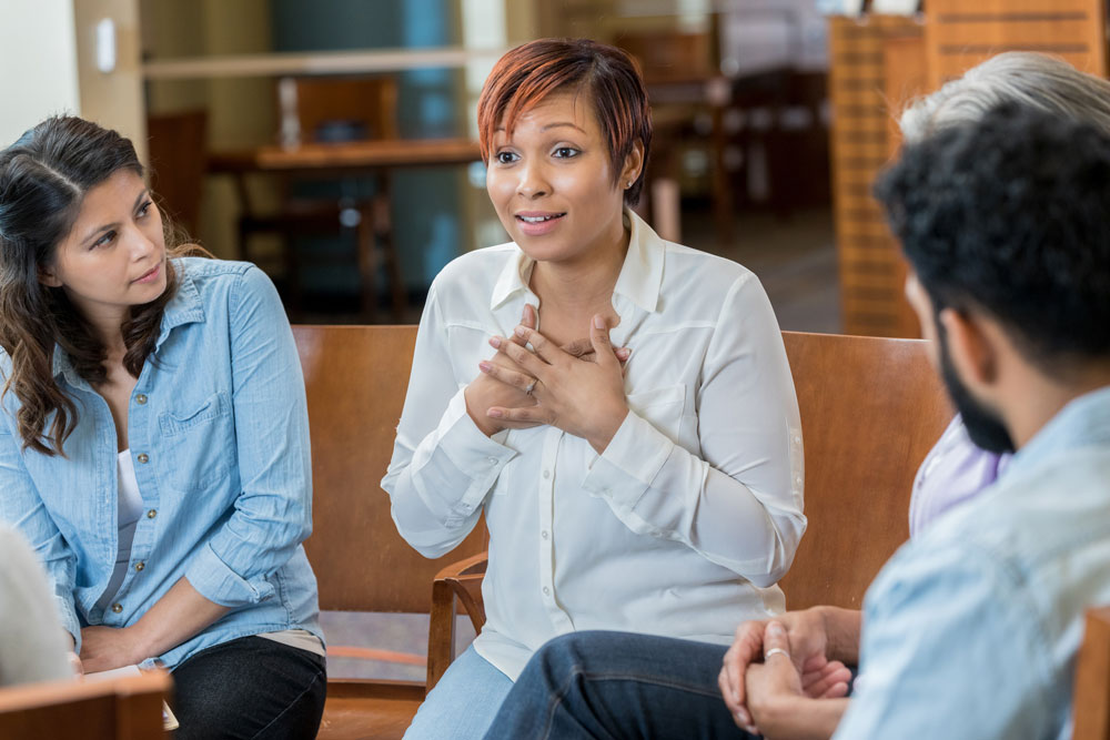 A woman sits in a circle with others, hands on her chest, speaking sincerely. Three people listen attentively, suggesting a support or group therapy session in a casual indoor setting.