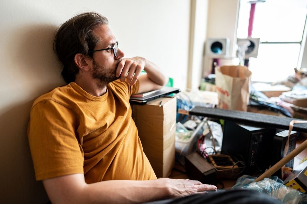 A man in glasses and a yellow shirt sits on the floor, leaning against a wall with a thoughtful expression. He is surrounded by clutter, including boxes, bags, and miscellaneous items in a messy room.