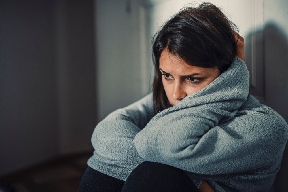 A woman in a gray sweater sits on the floor, hugging her knees and looking down with a worried, sad expression. She appears to be deep in thought or feeling anxious.