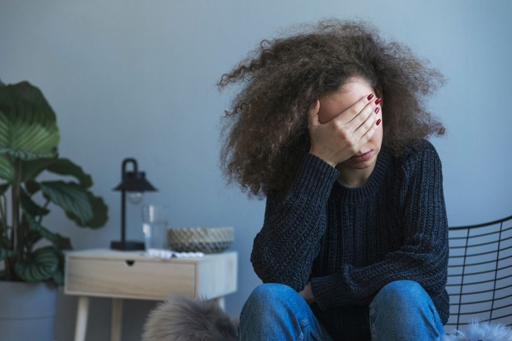 A person with curly hair sits indoors on a chair, wearing a dark sweater and jeans, with one hand covering their face, appearing stressed or upset. A small table with a lamp and plant is in the background.