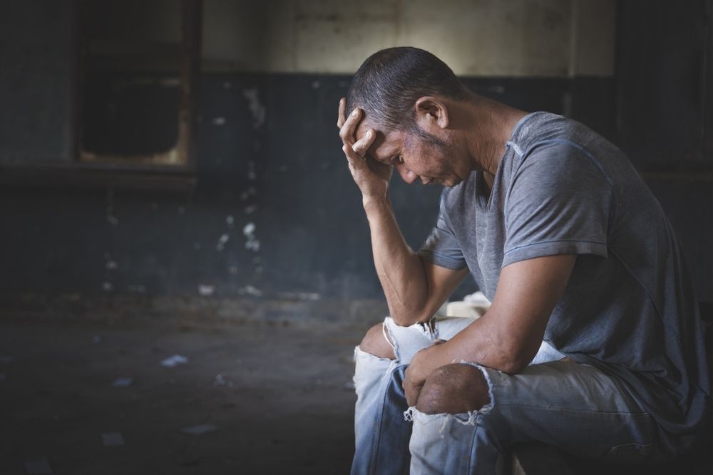 A man sits on a bench indoors, hunched over with his head resting on one hand, looking distressed. He is wearing a gray t-shirt and ripped jeans. The room appears dim and worn.