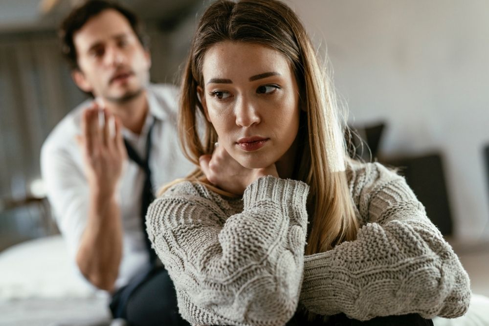 A woman with long hair sits with her arms crossed, looking upset. In the blurred background, a man gestures with his hand as if speaking to her, suggesting a tense conversation or argument.