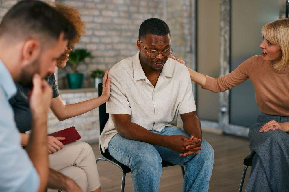 A man sits with his head down, looking upset, while three people around him offer comfort and support by touching his shoulder and back in a group setting.