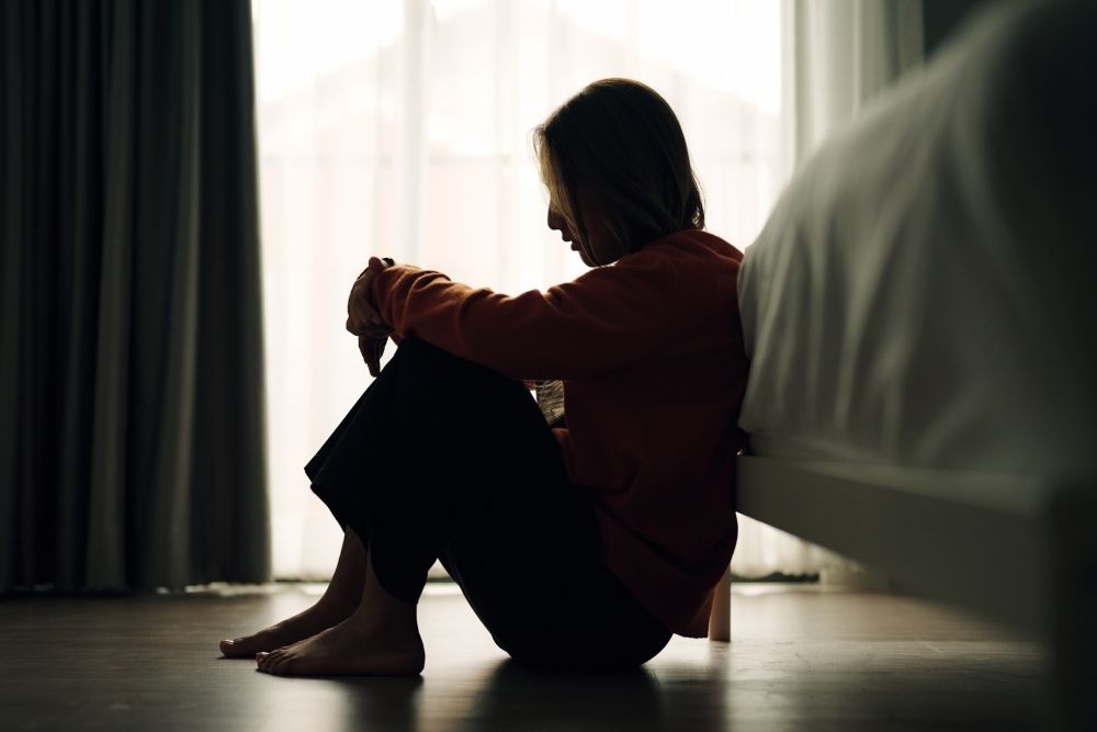 A person sits on the floor beside a bed, hugging their knees, with their head down in a dimly lit room. Light filters through sheer curtains in the background, creating a somber and contemplative mood.