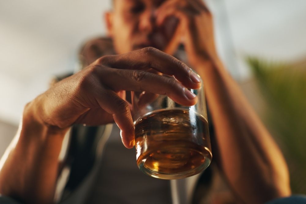 A close-up of a person holding a glass of whiskey with ice, their face blurred in the background, appearing contemplative or distressed.