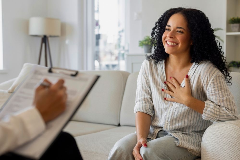 A woman sits on a couch, smiling and holding her hand to her chest, speaking with a person who is taking notes on a clipboard, suggesting a therapy or counseling session in a cozy, well-lit room.