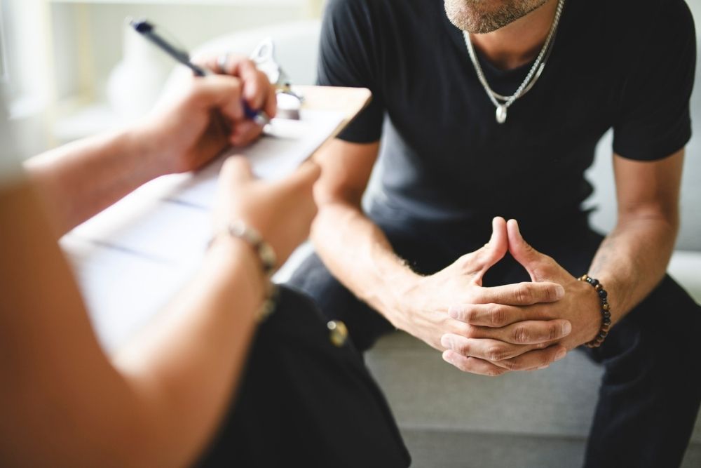 A person in a black shirt sits on a gray couch with hands clasped, while another person nearby writes on a clipboard, suggesting a counseling or therapy session.
