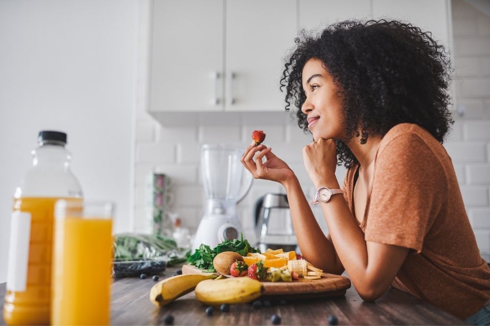 A woman with curly hair smiles thoughtfully while eating a strawberry at a kitchen counter with various fresh fruits, a glass and bottle of orange juice, and a blender in the background.