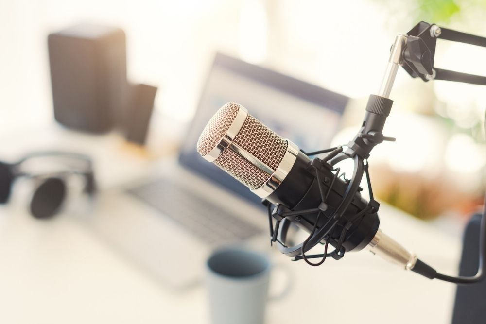 A close-up of a studio microphone on a boom arm in front of a blurred laptop, headphones, and a coffee mug on a desk, suggesting a podcast or recording setup.