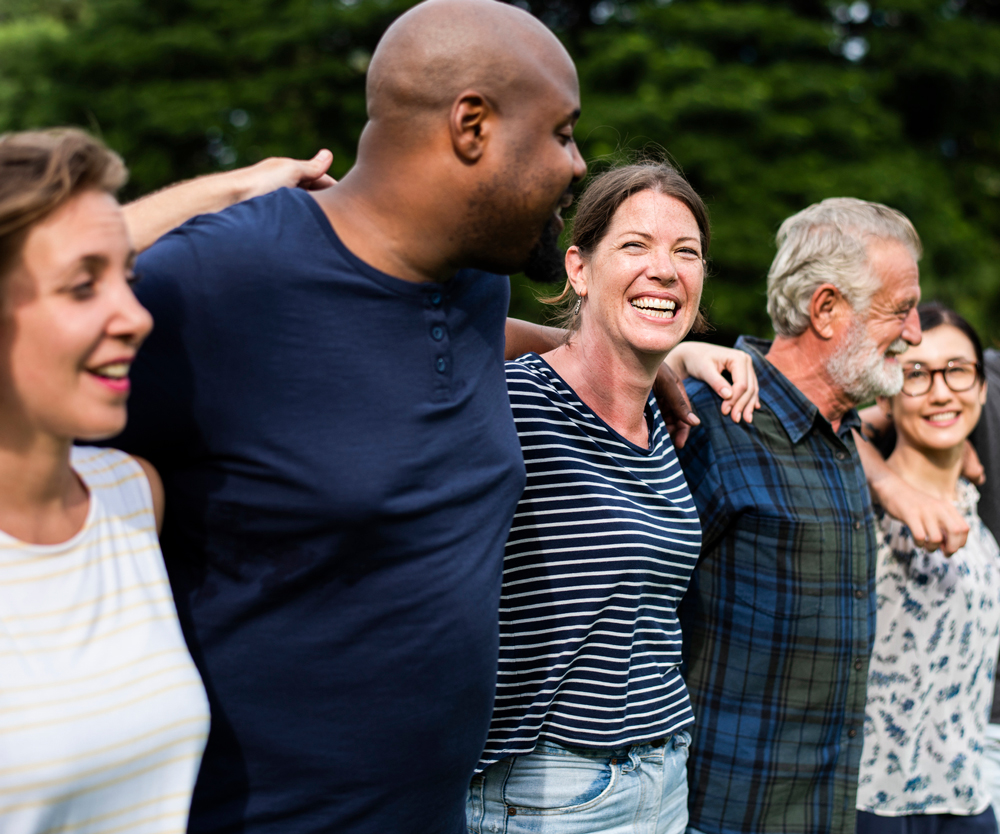 A group of five people standing outdoors with arms around each other, smiling and laughing. The background features green trees and grass, suggesting a park or garden setting.