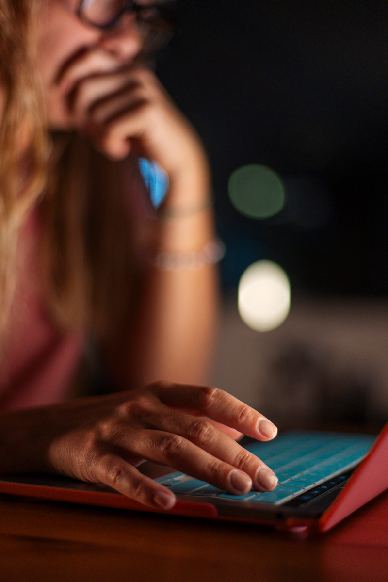 A person sits in low light, their face partially visible and hand resting on a laptop keyboard, appearing focused and thoughtful as they look at the screen.