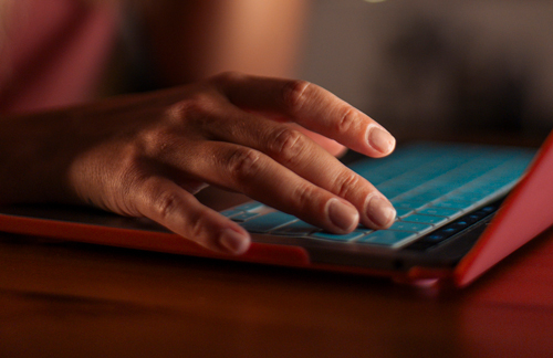 A close-up of a persons hand typing on a laptop keyboard with a blue silicone cover, illuminated by warm indoor lighting.