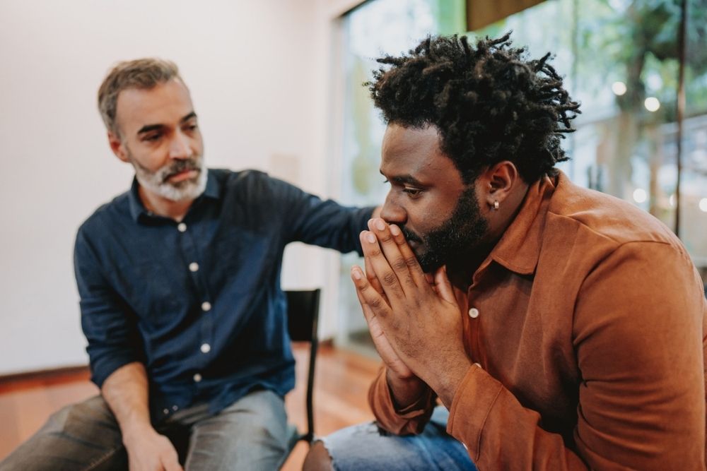 Two men sit indoors; one appears concerned with hands clasped near his face, while the other offers comfort by placing a hand on his shoulder.