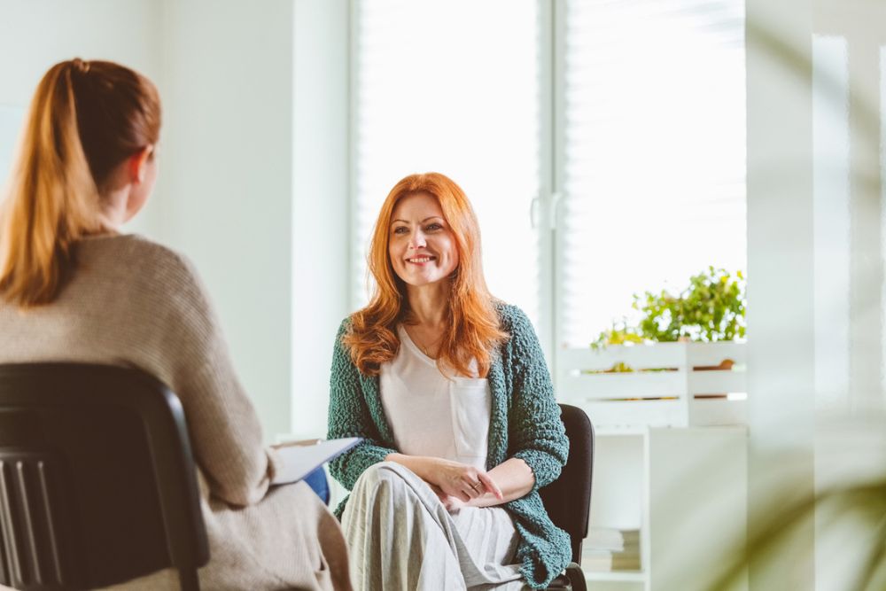 Two women sit facing each other in a bright room. One is smiling and appears relaxed, while the other takes notes. Sunlight streams through a window with plants on the windowsill.