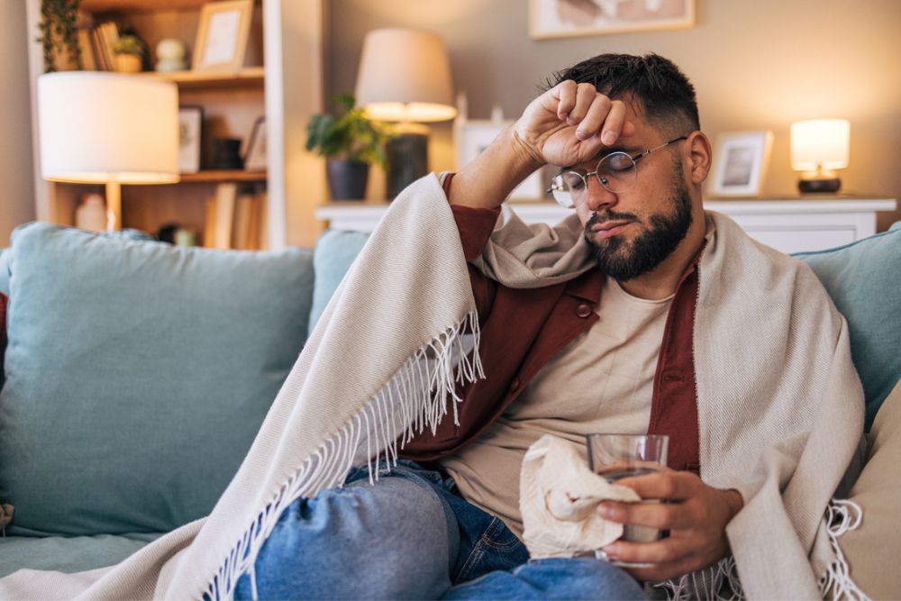 A man sits on a couch wrapped in a blanket, holding a glass of water and a tissue, with his hand on his forehead. He appears unwell and tired, in a cozy living room setting.