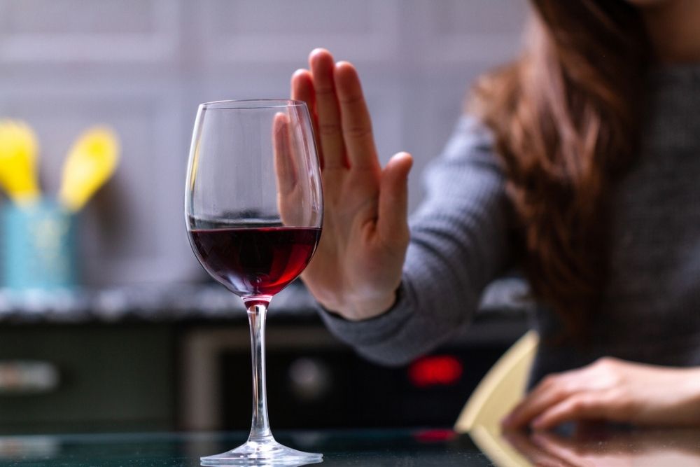 A person with long brown hair holds up a hand to reject a glass of red wine on a table, signaling refusal or abstaining from alcohol.