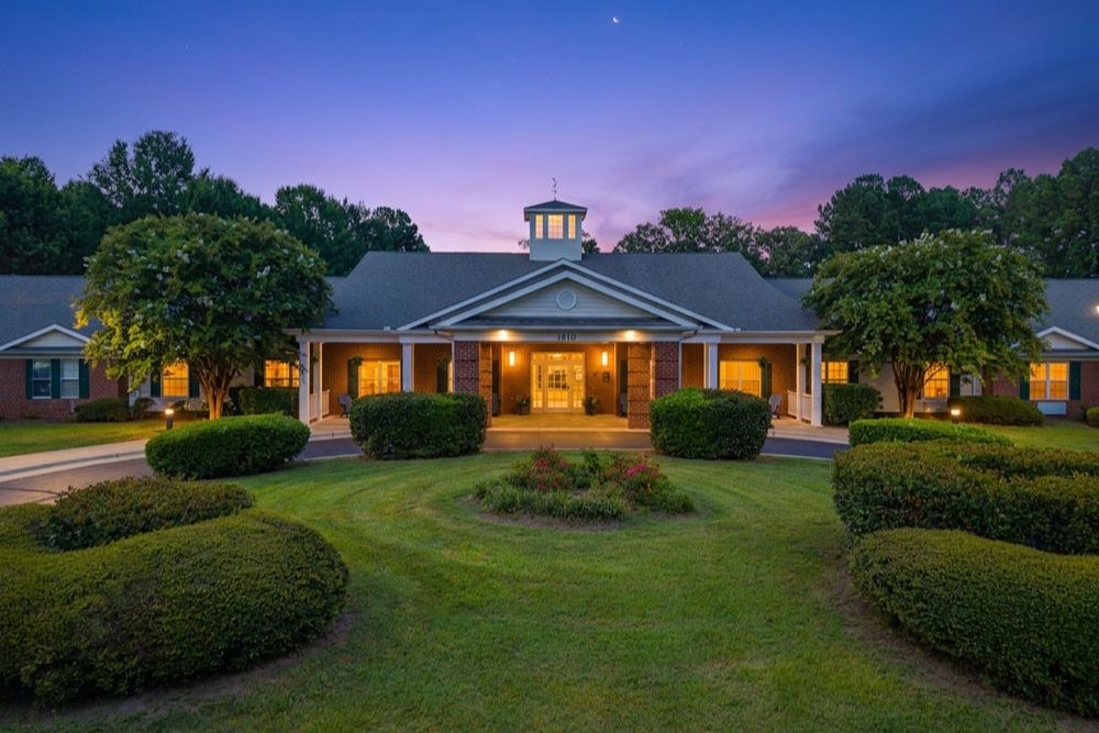A large, single-story building with a white entrance and columns is surrounded by neatly trimmed shrubs and a circular driveway, set against a vibrant night sky with trees in the background.