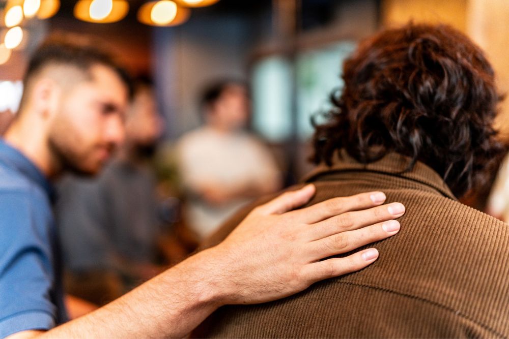 A person places a comforting hand on another persons shoulder, offering support. The background is blurred, showing other people and warm lights.