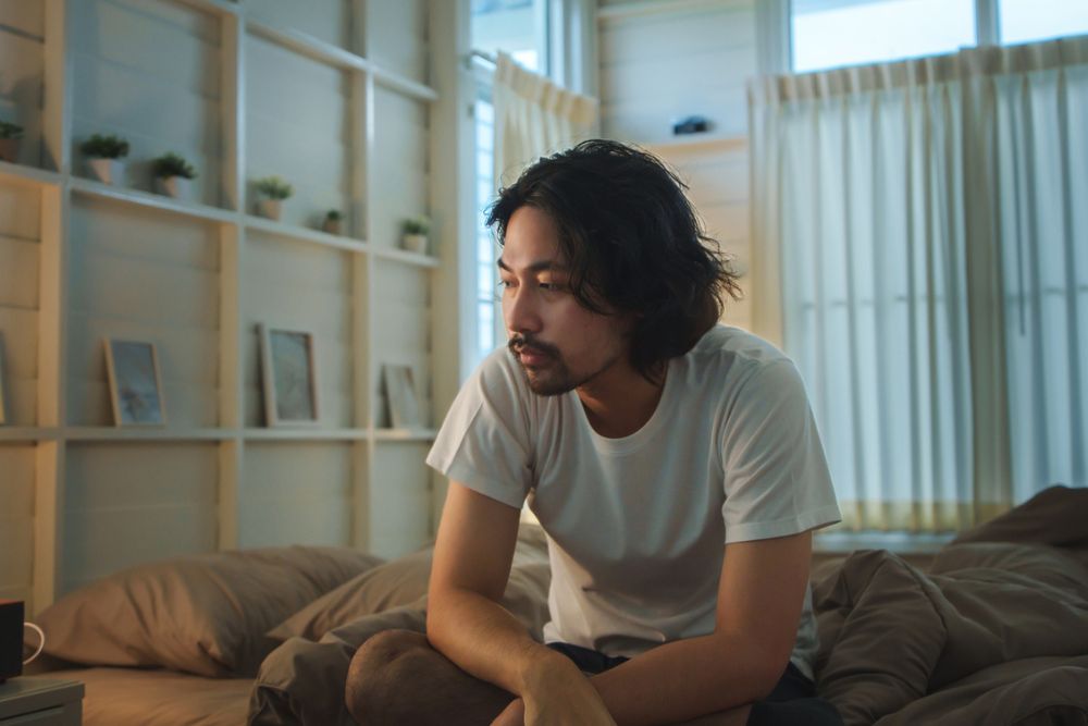 A man with medium-length hair and a beard sits on a bed in a softly lit bedroom, looking thoughtful. Shelves with plants and framed pictures decorate the wall behind him. Light filters through the curtains.