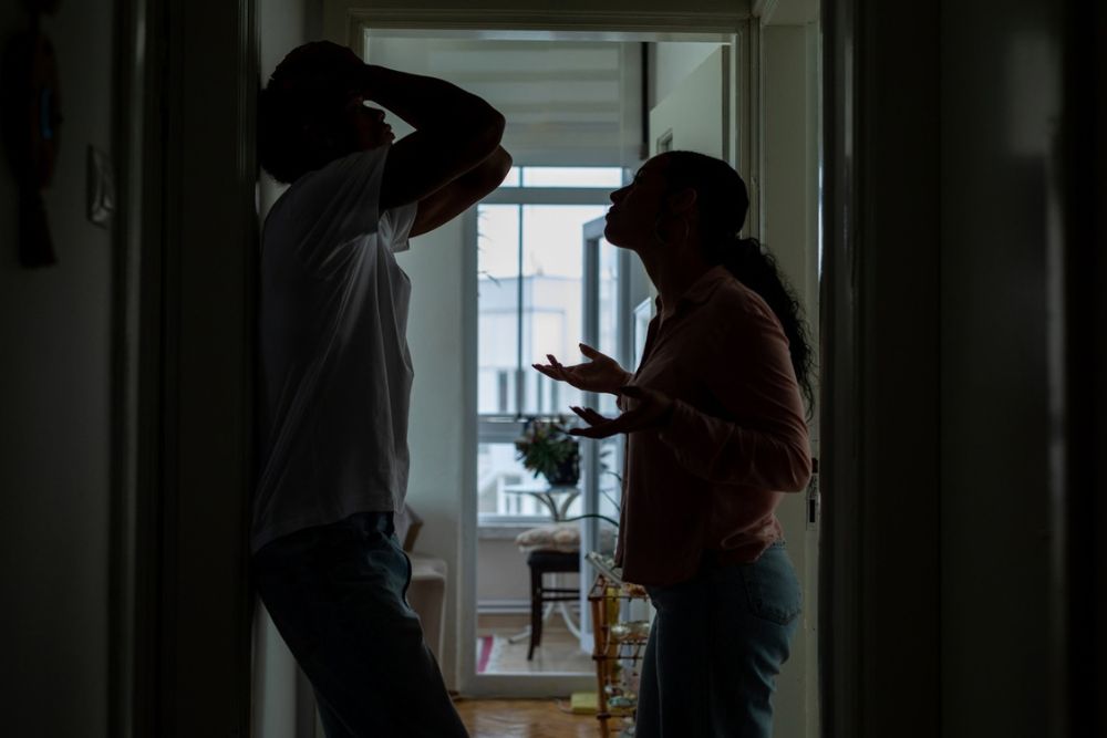Silhouetted man and woman stand in a hallway, appearing to argue. The man leans against the doorframe with his hand on his head, while the woman gestures with her hands. Daylight streams in from a window behind them.